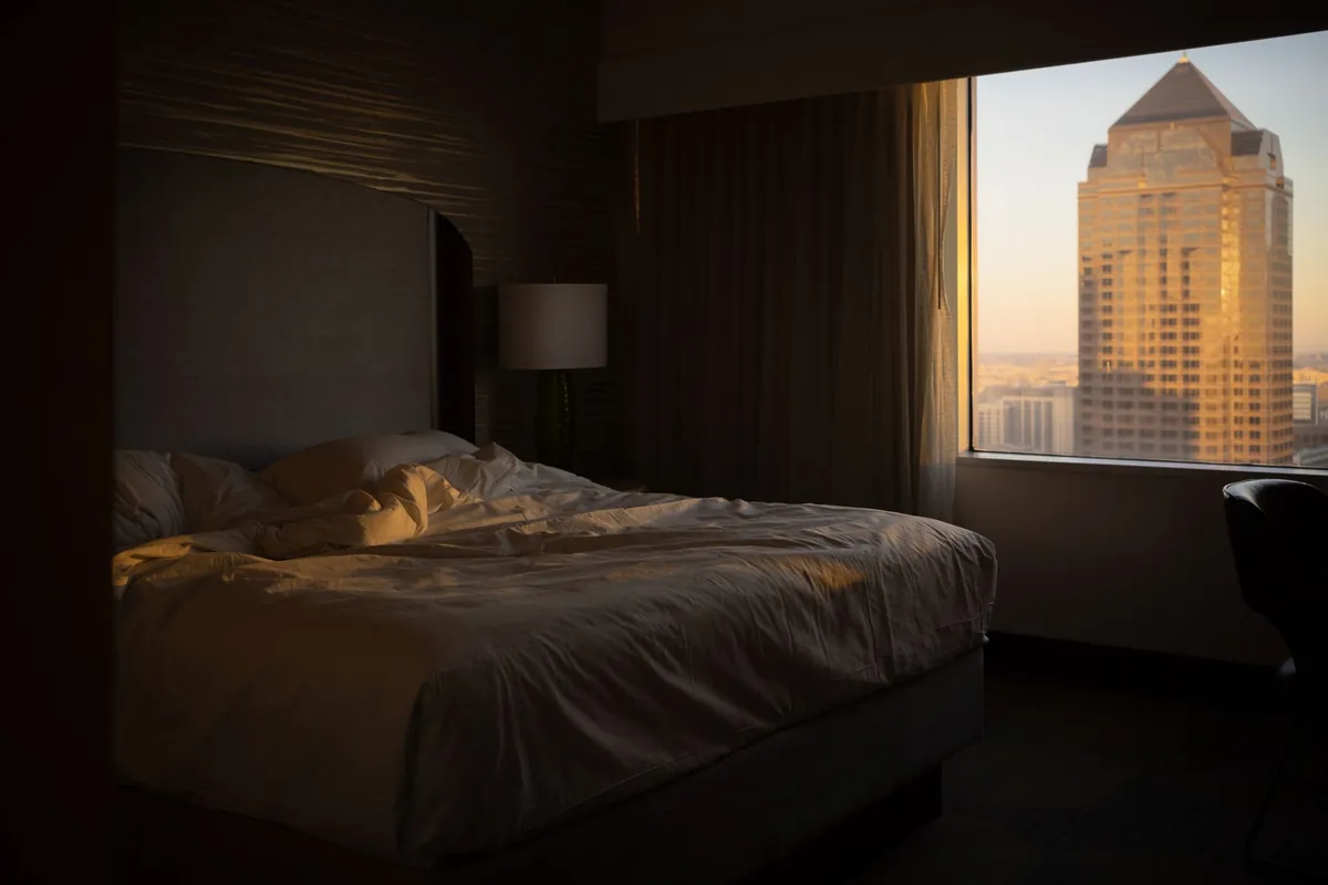 A made bed in a dim hotel room with a city view through the window