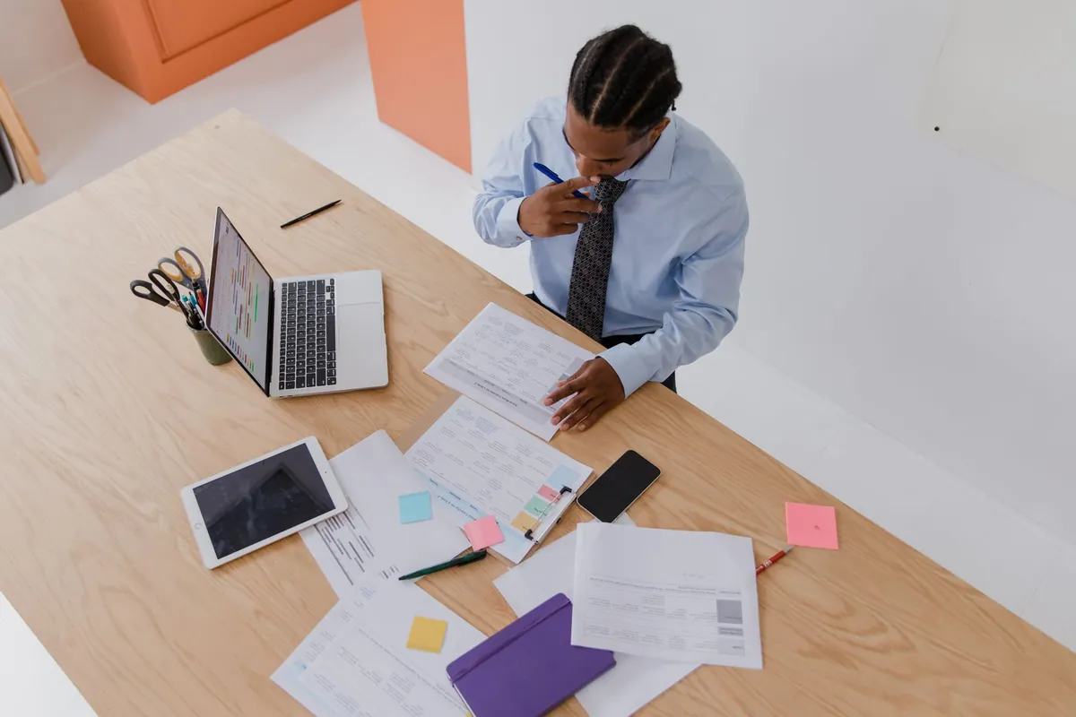 A man at a desk working through printed documents beside a tablet and a laptop