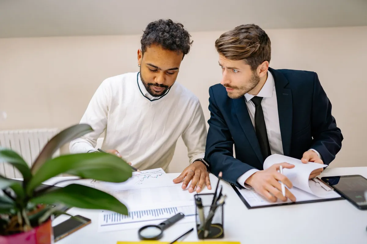 Two people talking across a desk with documents and a laptop between them