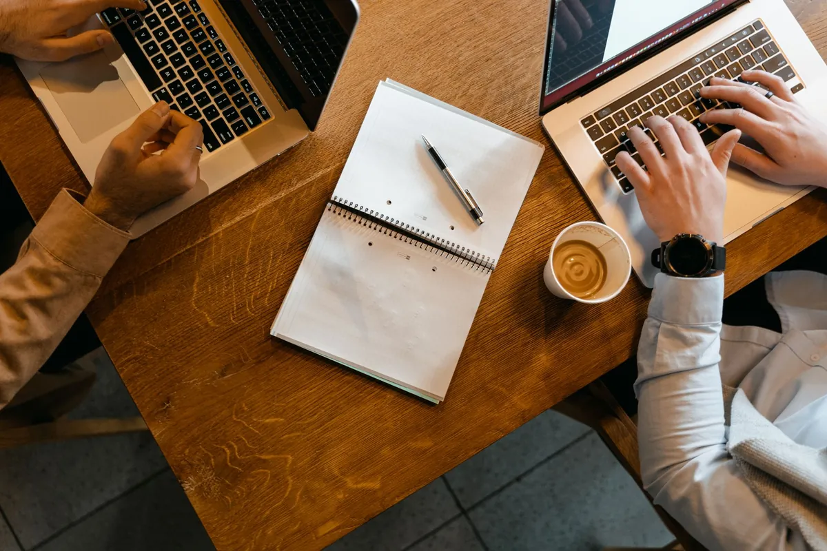 Overhead view of two people working at laptops with notes and coffee on a wooden table