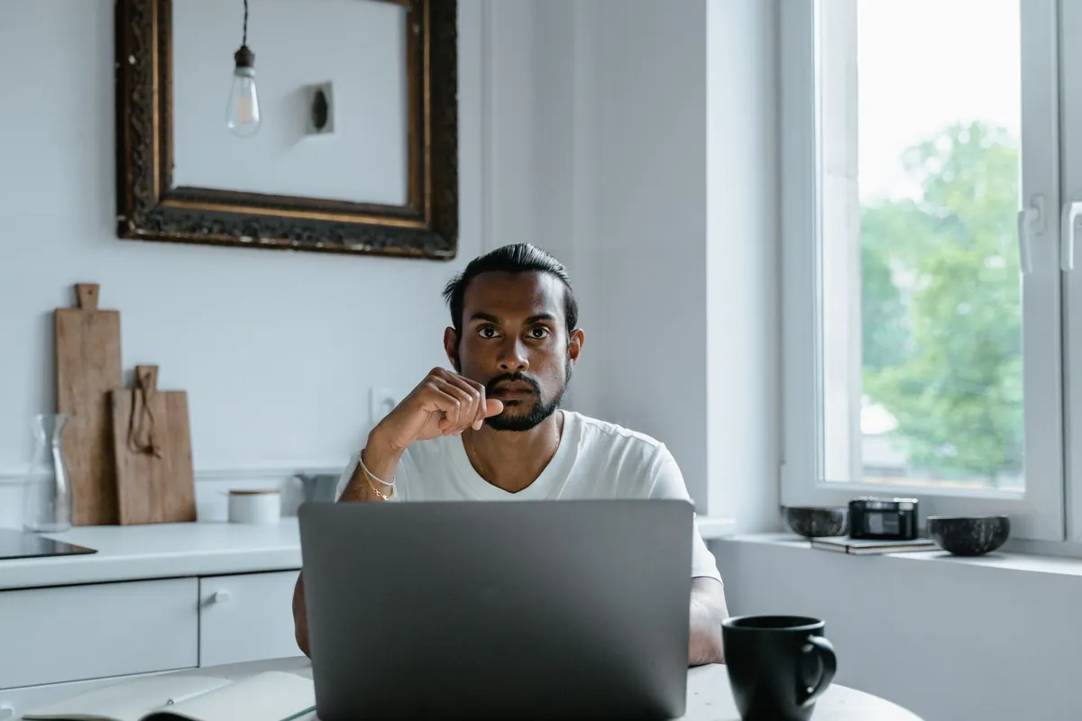 A man sitting with a laptop at a kitchen counter, thinking