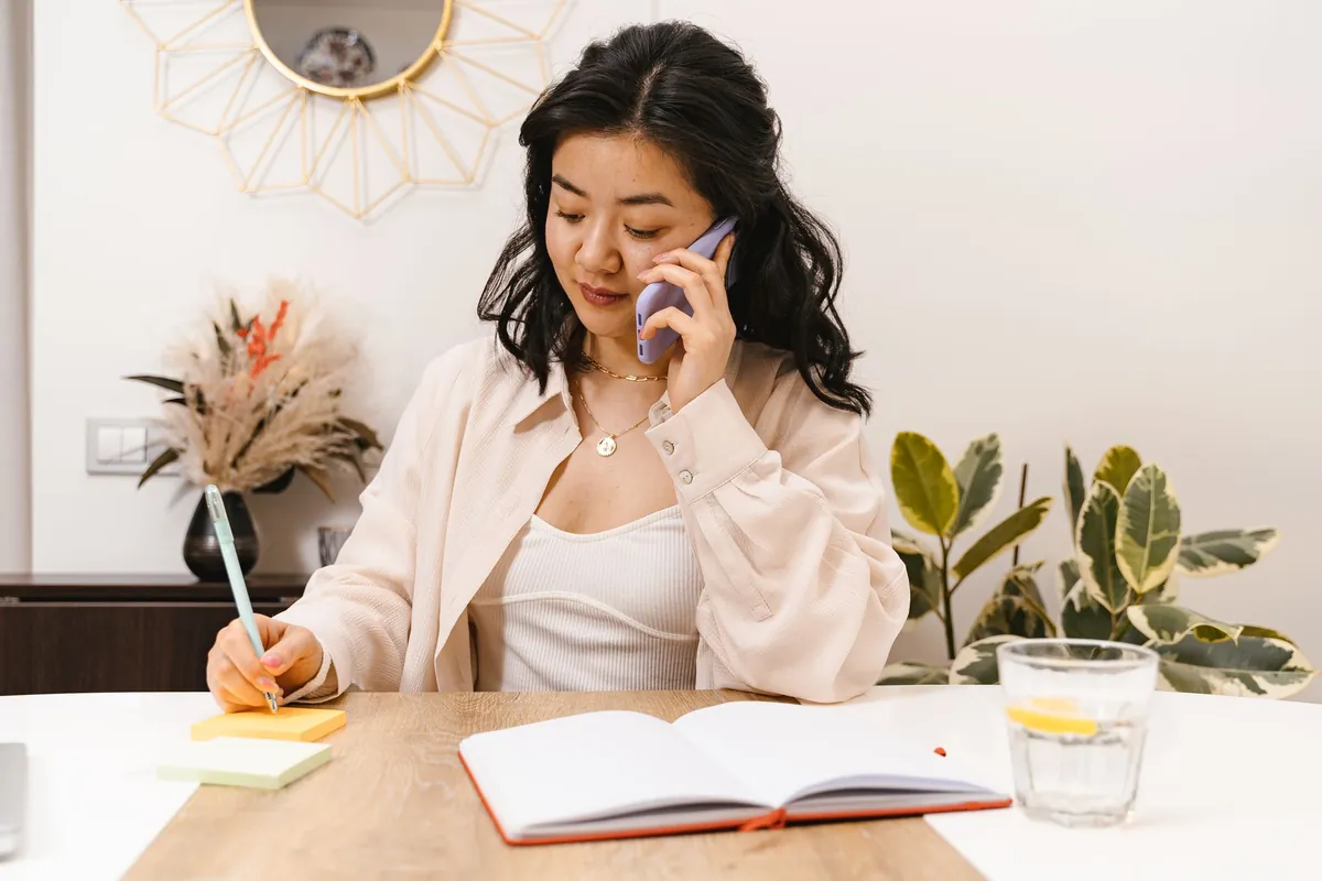 A woman at a light wooden table holding a phone to her ear with one hand and a pencil in the other, an open blank notebook and a glass of water in front of her.