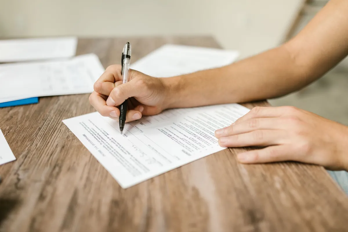 Two hands at a wooden table, one holding a pen over a printed form, with more loose sheets behind it.