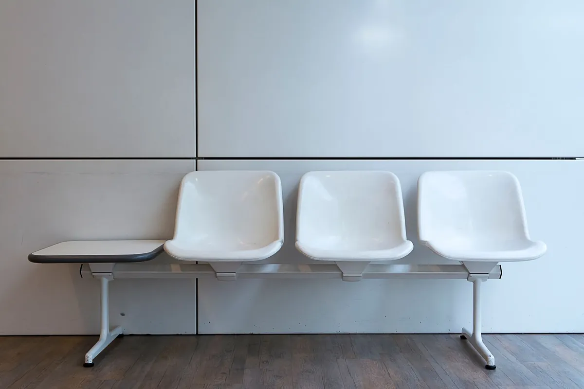 Three empty white moulded chairs on a metal beam bench against a plain white wall, with a small side shelf and a wooden floor.