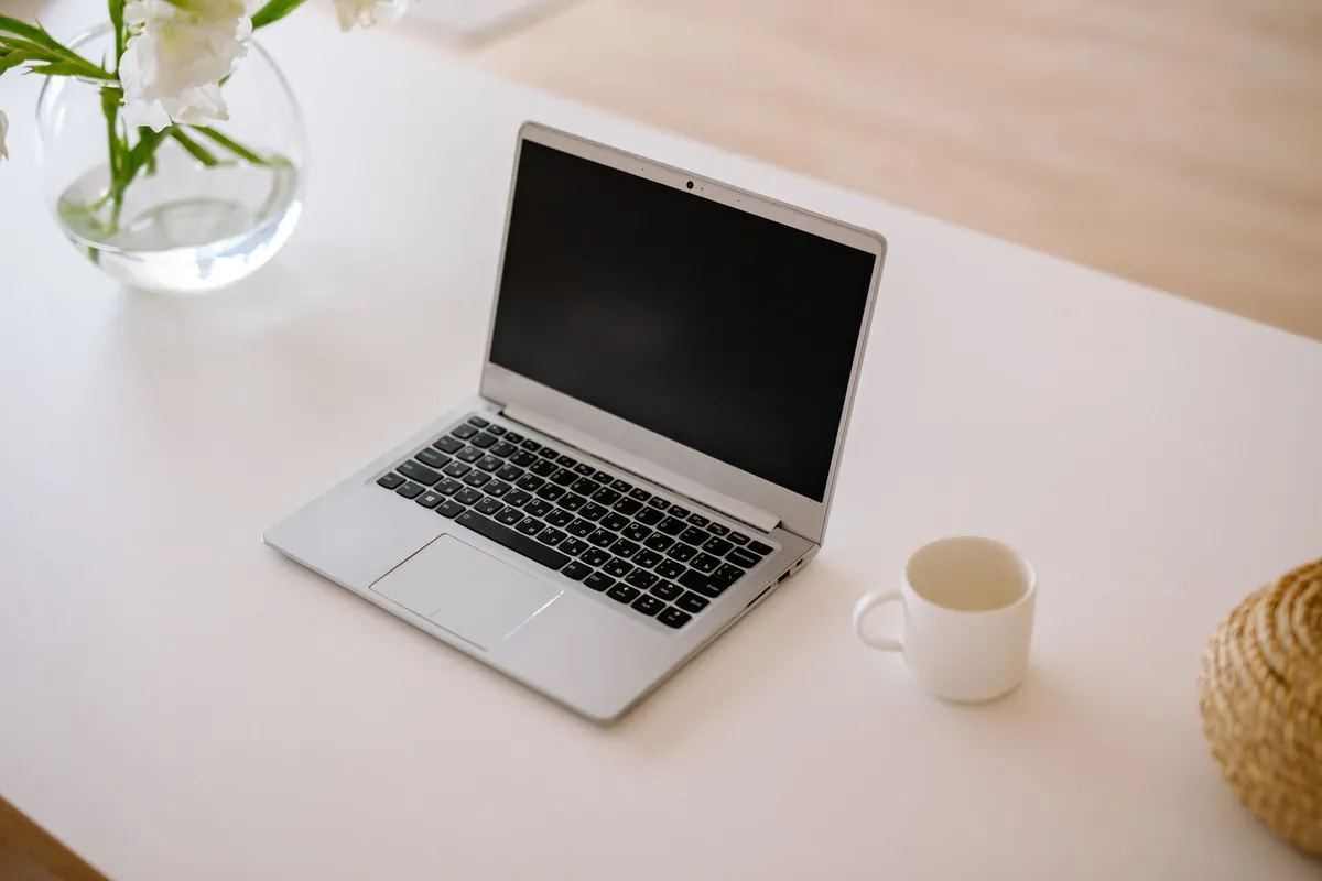 A single open silver laptop with a dark blank screen on a white table, beside a plain white mug and a glass vase of white flowers.