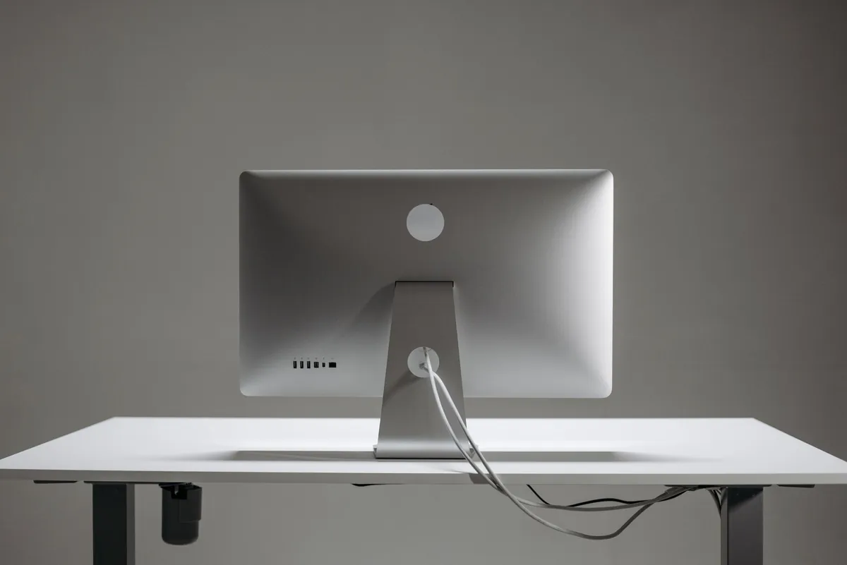 The back of an aluminium all-in-one monitor on a white height-adjustable desk against a grey wall, its cable trailing down.