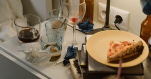 A cluttered bedside table after a stay: glasses, a wine glass, crumpled tissues and a plate with pizza