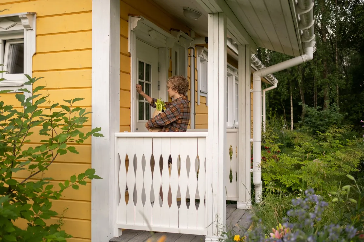 A person knocking at the door of a yellow cottage porch