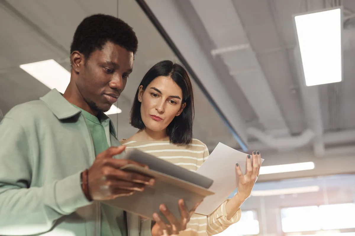 Two people examining a tablet and printed pages together in a bright office