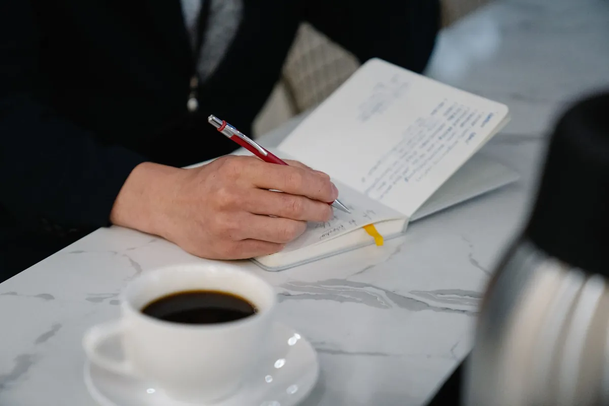 A hand writing notes in a journal beside a cup of black coffee