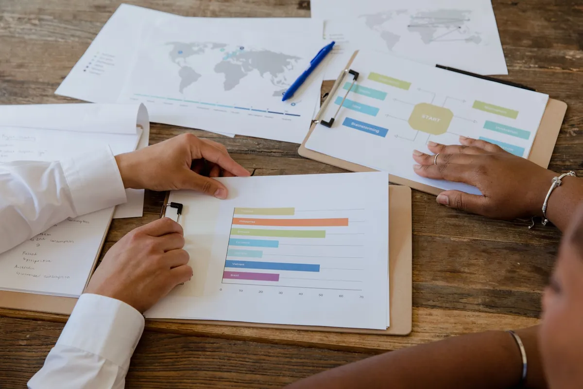 Looking down on a wooden table where two people's hands rest on printed charts - a world map, a flow diagram and a coloured bar chart - with a blue pen lying across them.
