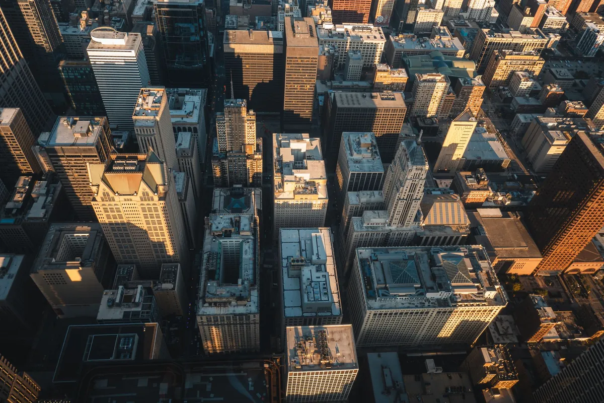 A steep aerial view straight down onto a downtown grid of office towers and rooftops, lit low from one side.