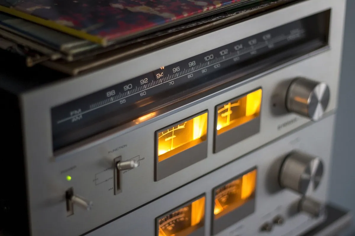 A close-up of a vintage hi-fi tuner with an illuminated frequency scale, two orange-lit meters and large brushed metal control knobs, with record sleeves stacked on top.