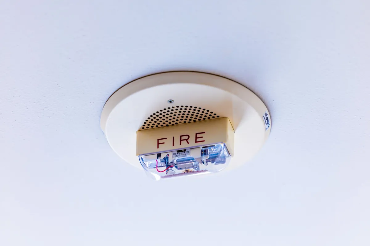 A round white ceiling-mounted alarm with a perforated grille, a small amber strobe housing and the word FIRE printed on it.