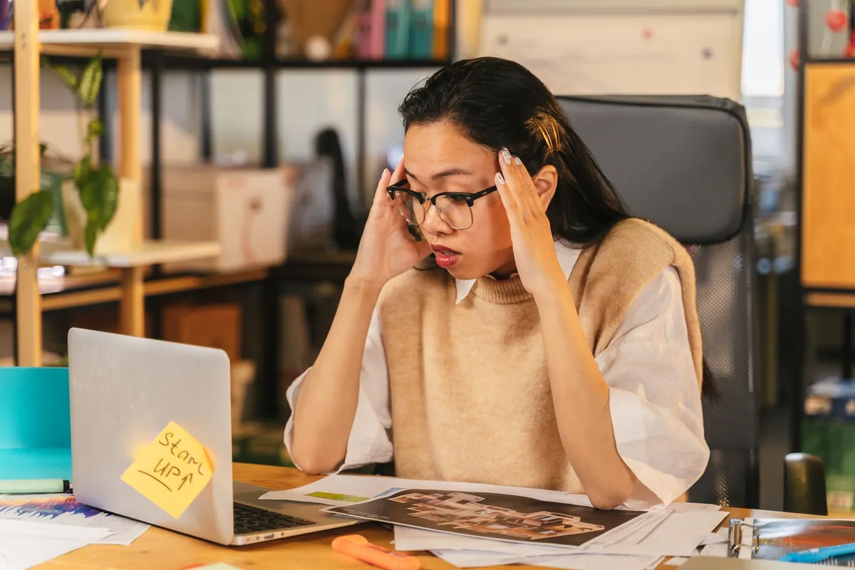 A person at a desk with a laptop and papers, rubbing their temples while reading