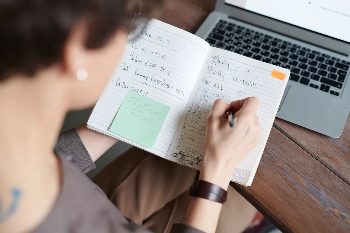 A person writing in a notebook beside a laptop on a wooden table