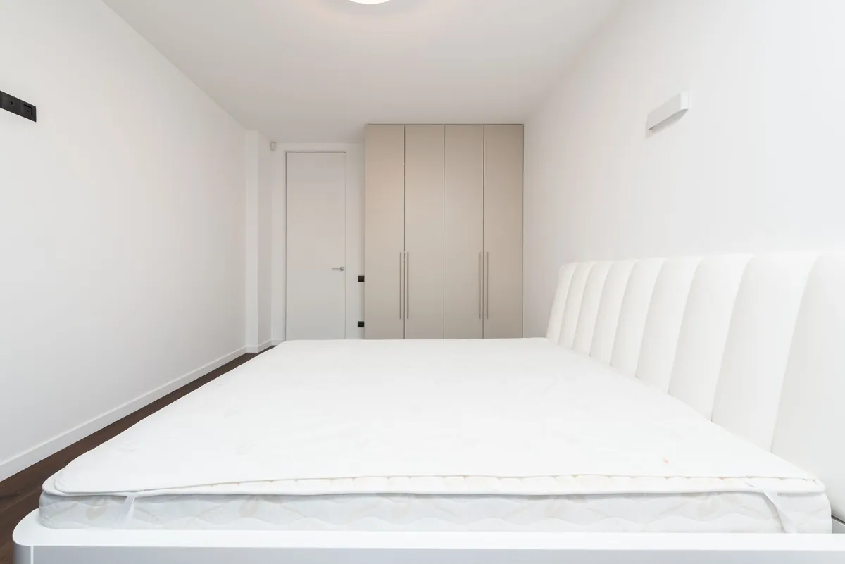 A bare white mattress on a bed frame in an empty white bedroom
