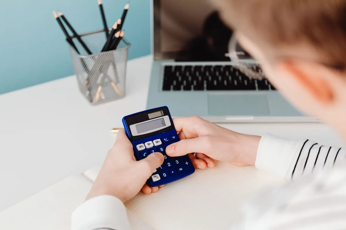 A person working out numbers on a blue calculator in front of a laptop