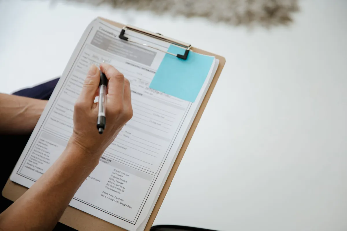 A hand filling in a printed form clipped to a clipboard, a blue sticky note beside it