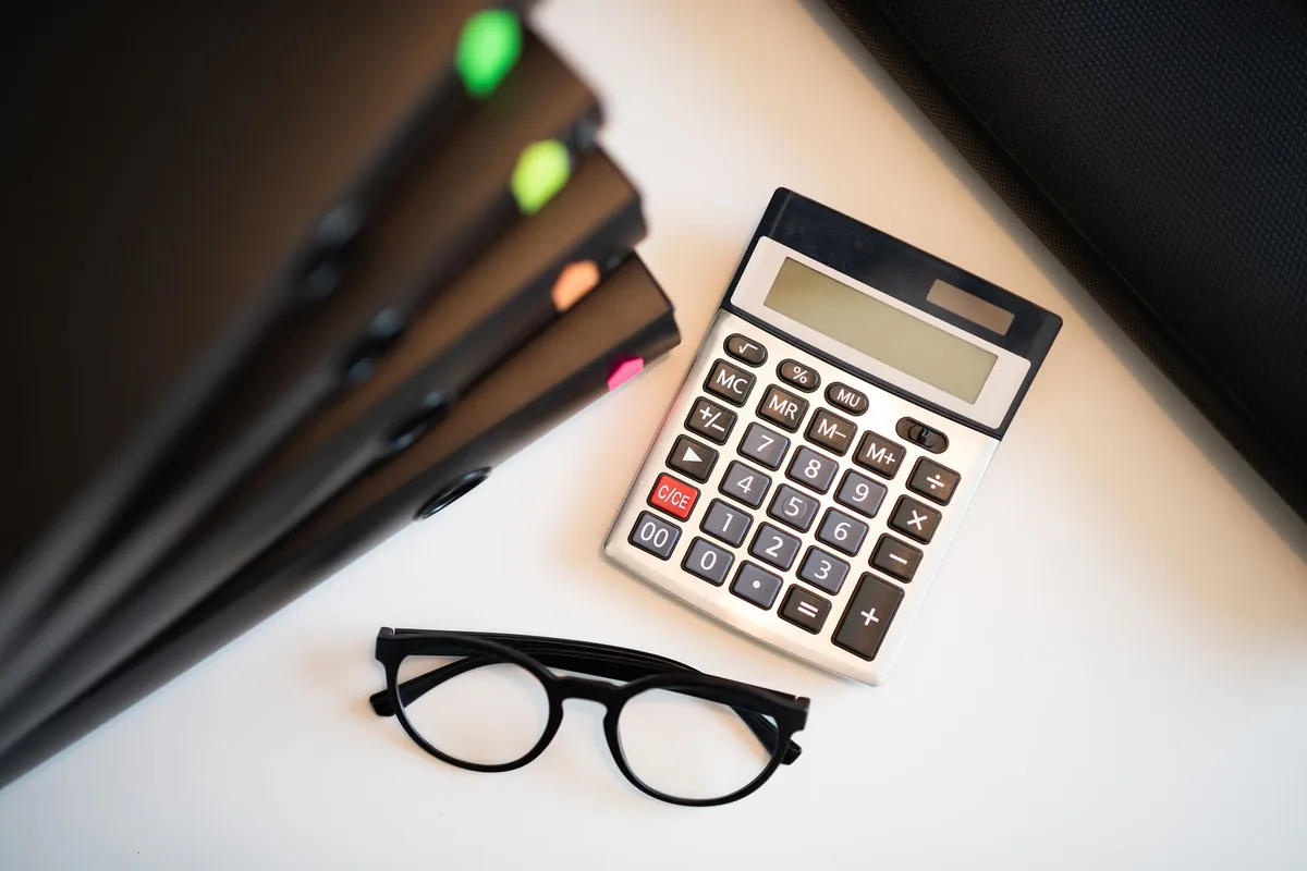 A white desk calculator lying beside a pair of glasses and a stack of dark folders