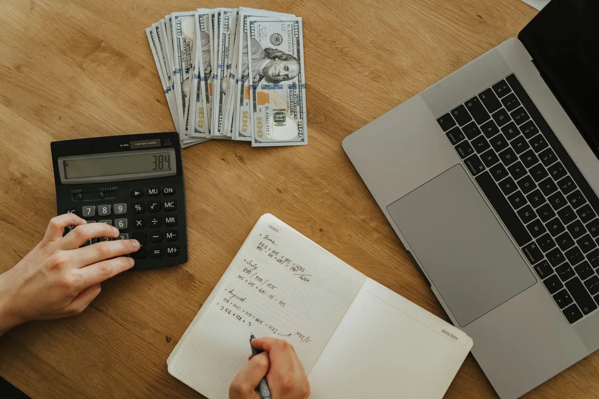Hands working through calculations in a notebook beside a calculator and a laptop, with cash on the desk