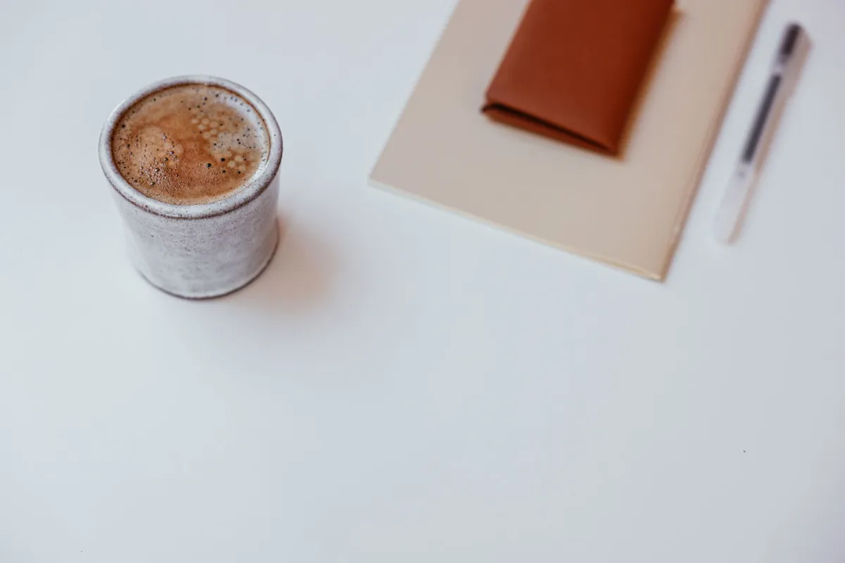 A cup of coffee, a closed notebook and a pen on a white desk