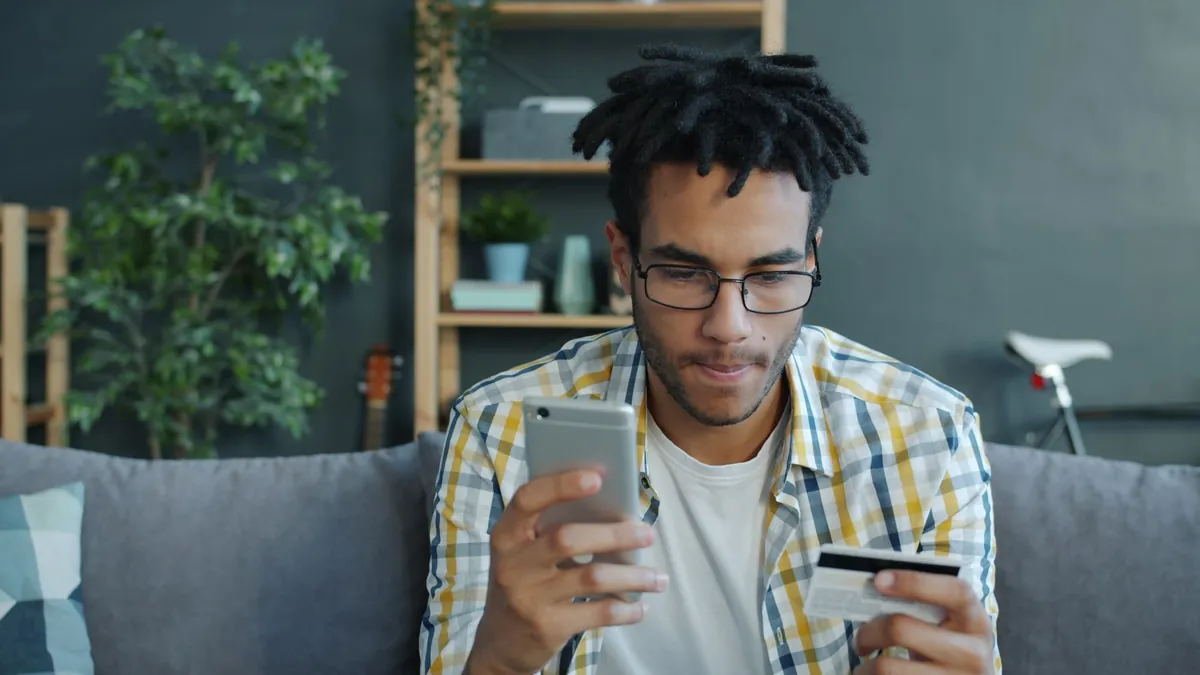 A man on a sofa holding a phone in one hand and a bank card, back side up, in the other