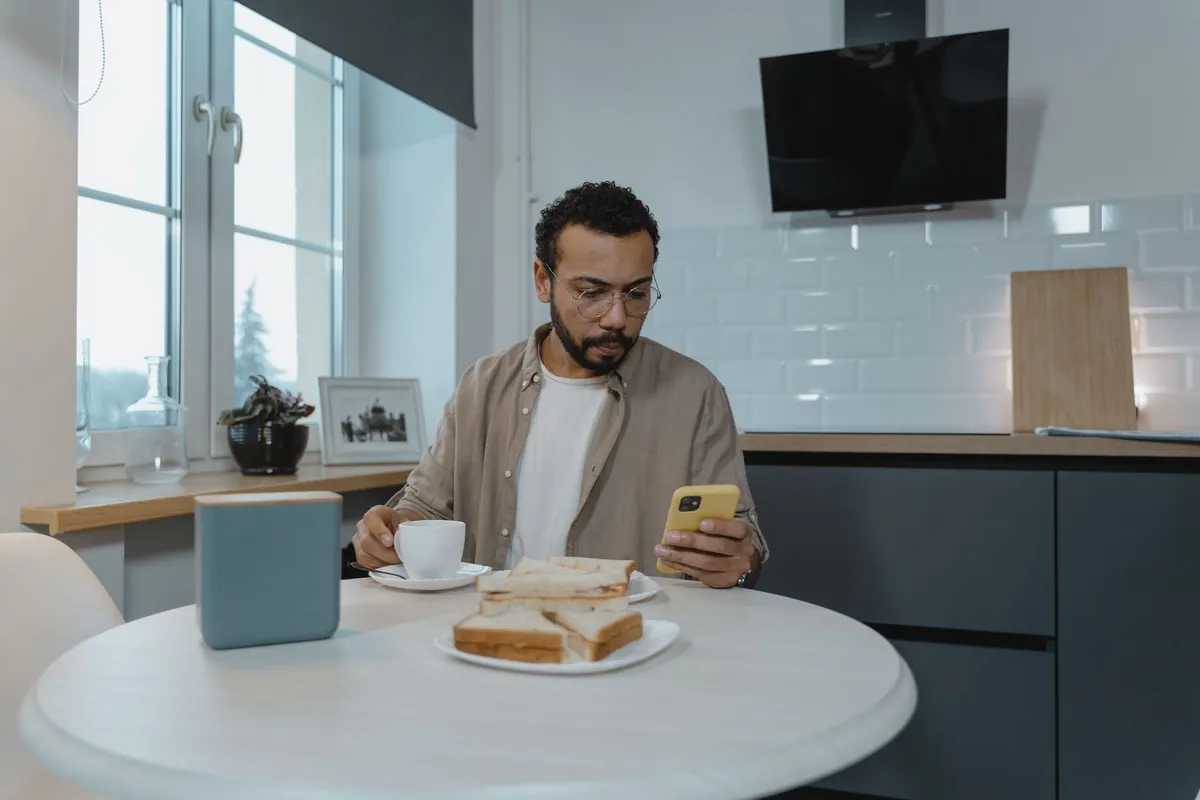 A man at a kitchen table reading a message on his phone next to a plate of sandwiches