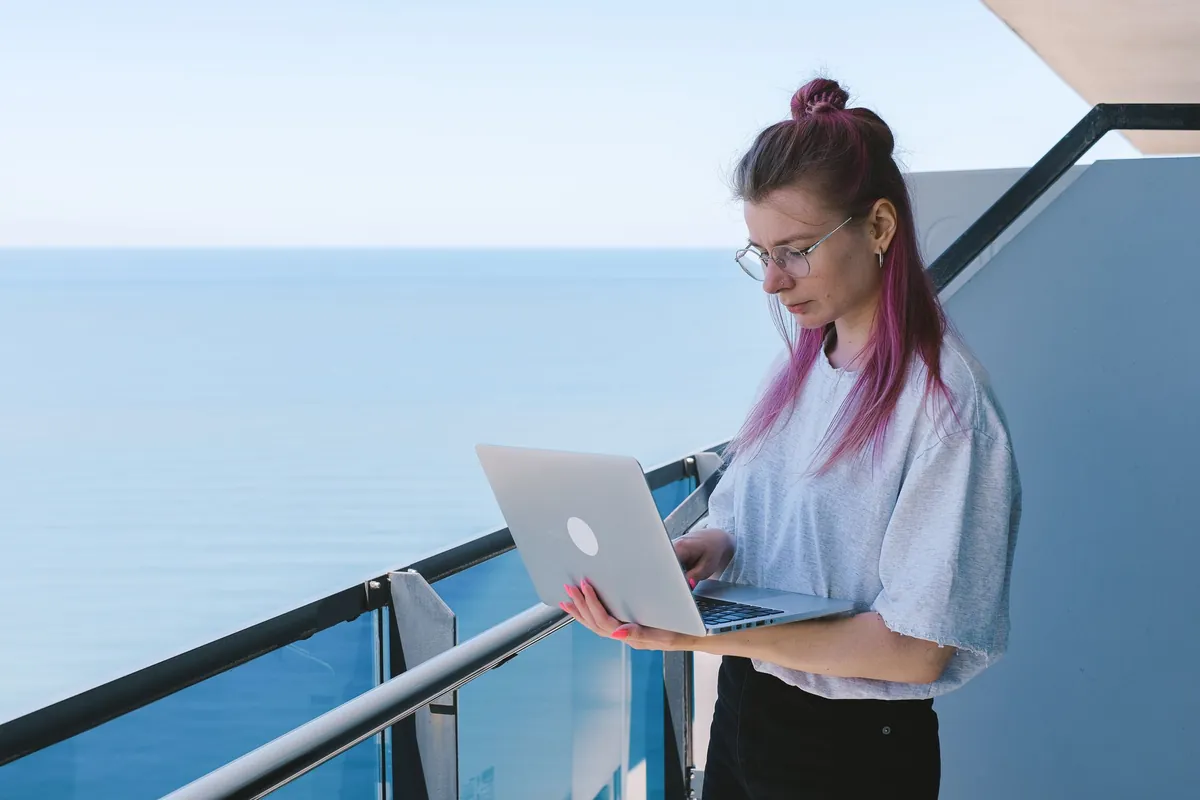 A host standing at a balcony railing with a laptop, the sea filling the frame behind her