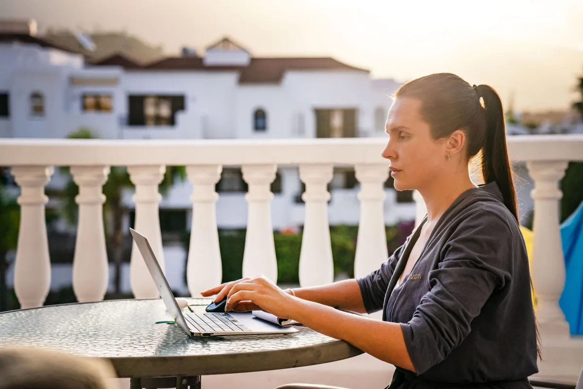 A woman working on a laptop at an outdoor table with white buildings behind her
