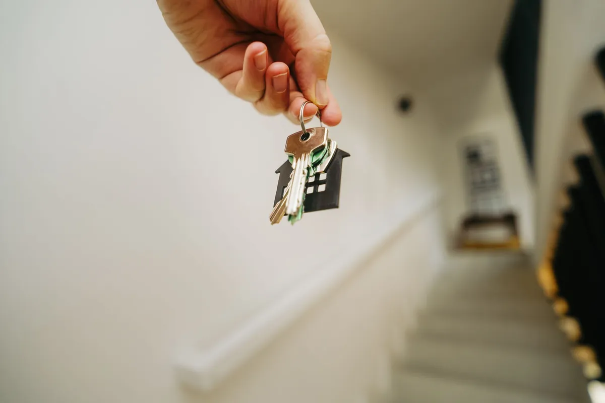 A hand holding a keyring with several keys and a small house charm on a stairwell landing