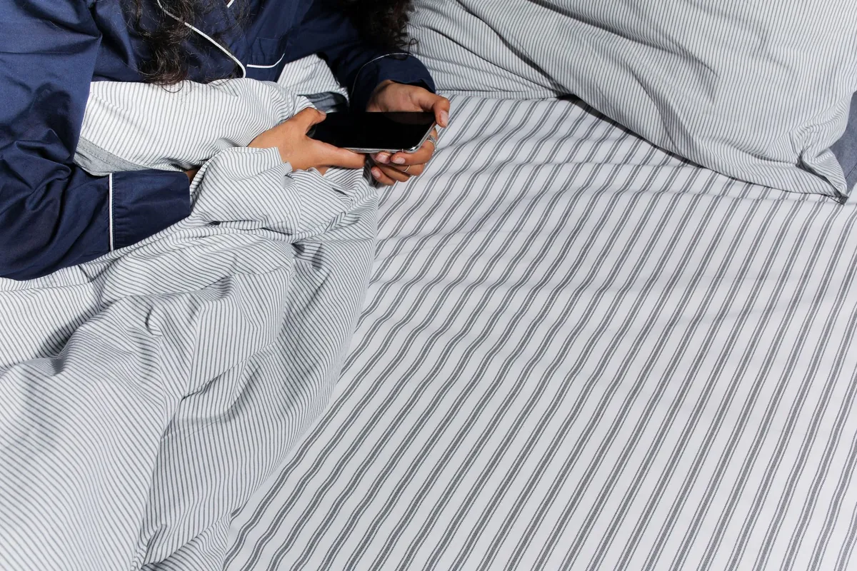 A person lying in bed under striped covers, holding a phone in the dark