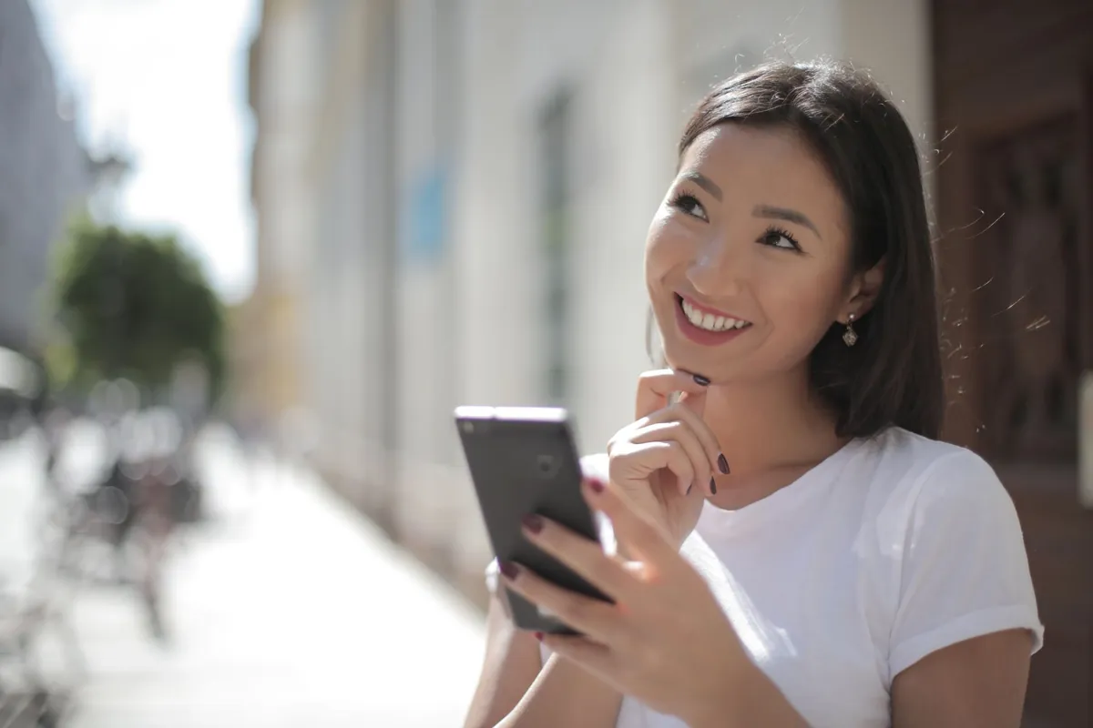 A woman standing outdoors looking at her phone with a thoughtful expression
