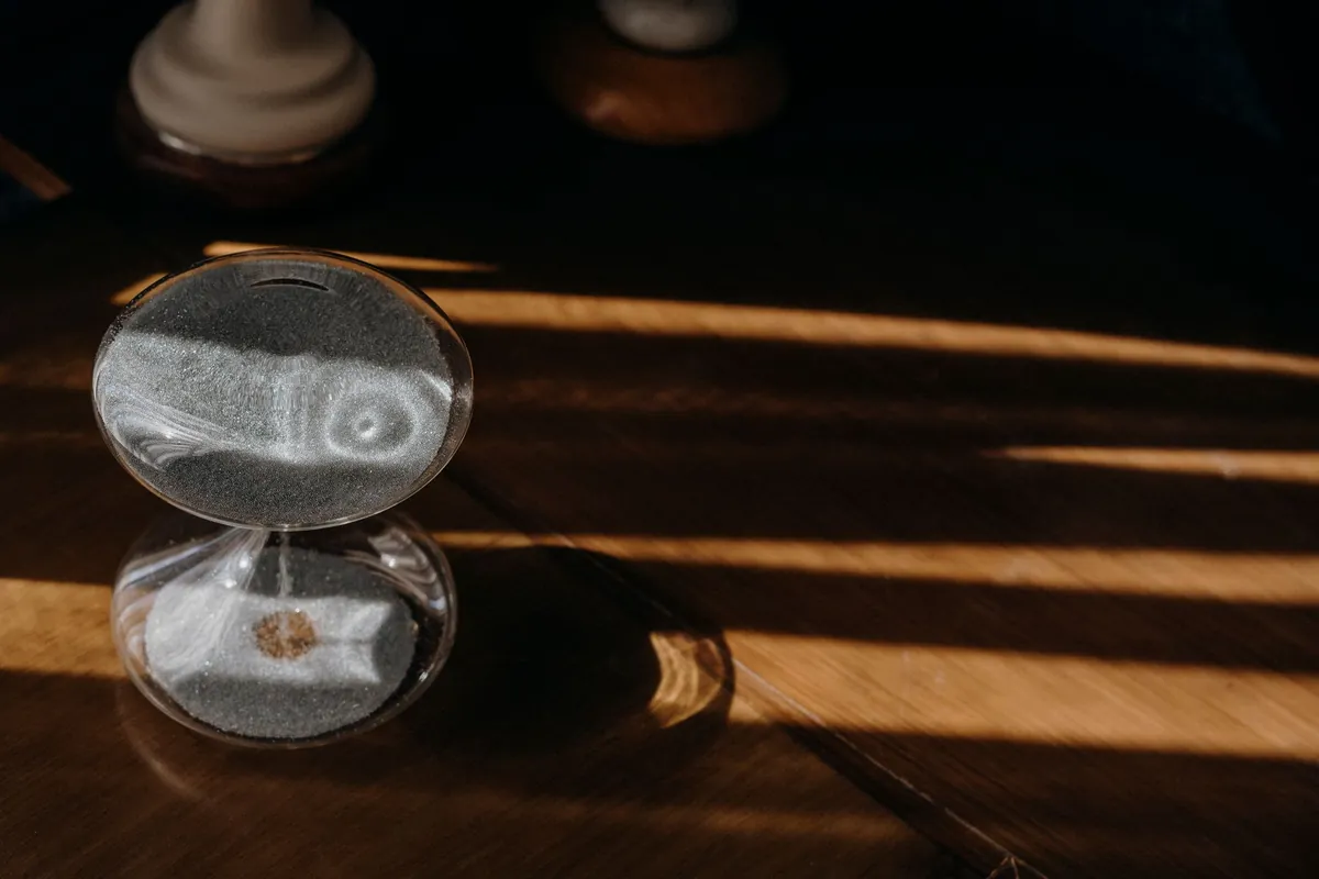 A small glass hourglass with fine white sand standing on a wooden table, striped by bars of morning sunlight.
