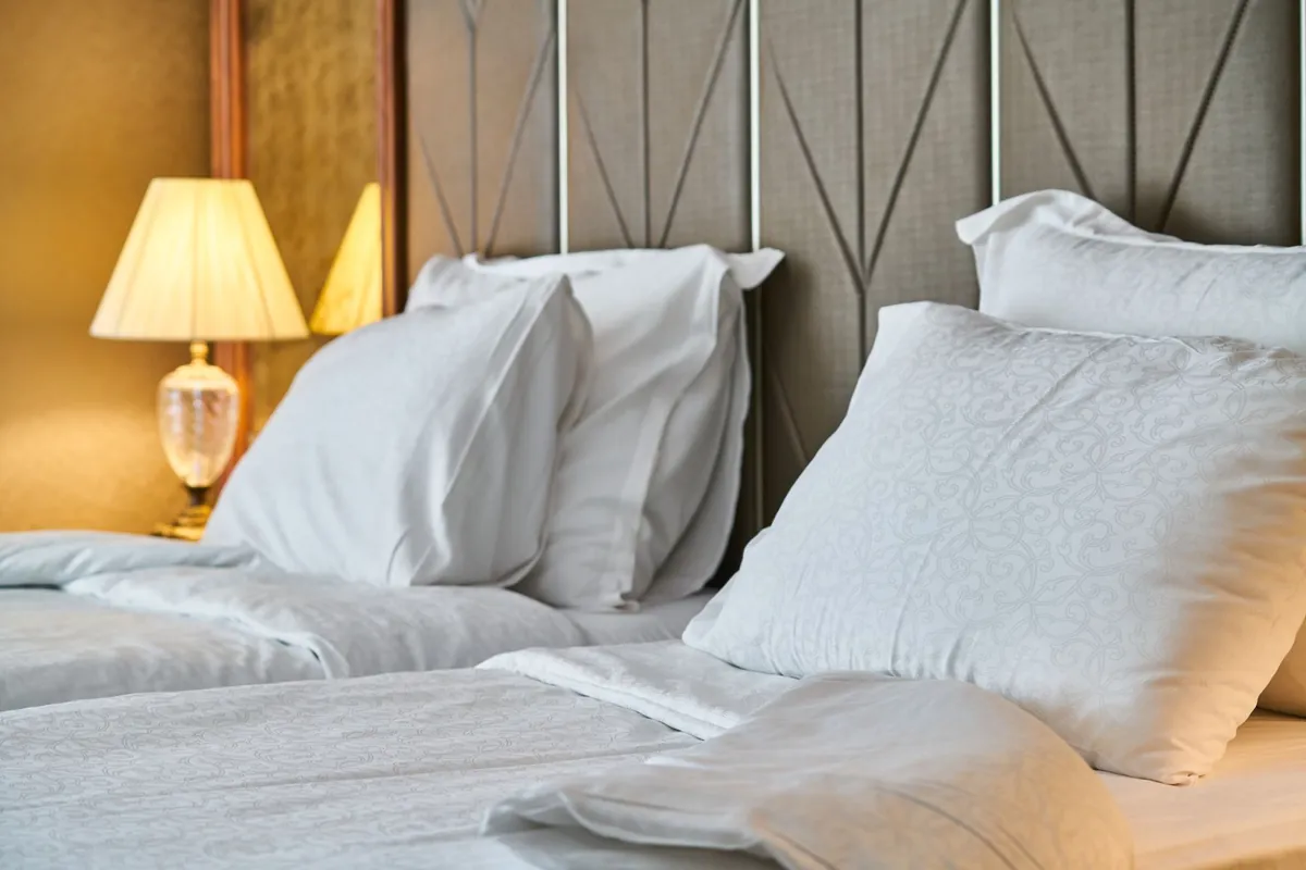 A made-up bed with crisp white linen and stacked pillows against an upholstered headboard, lit by a warm bedside lamp.