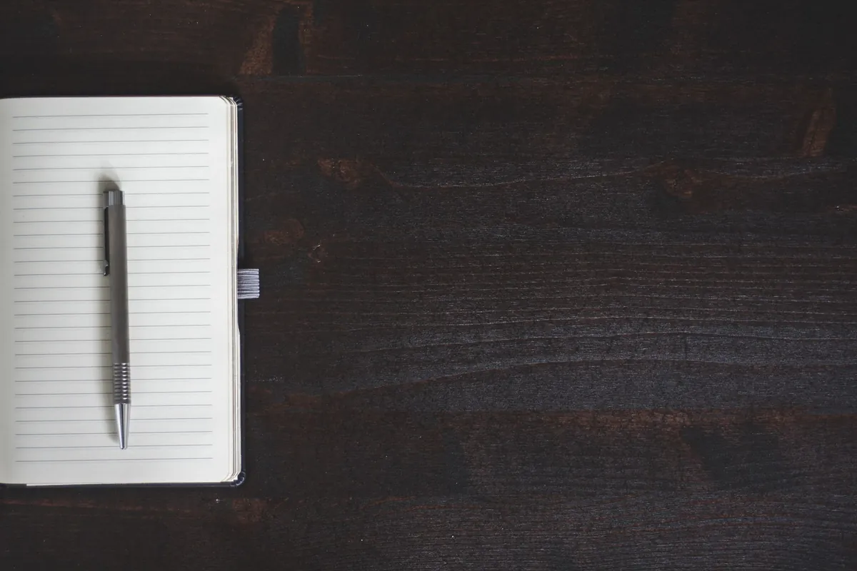 An open notebook with blank lined pages and a plain silver ballpoint pen lying on it, on a dark stained wooden desk.