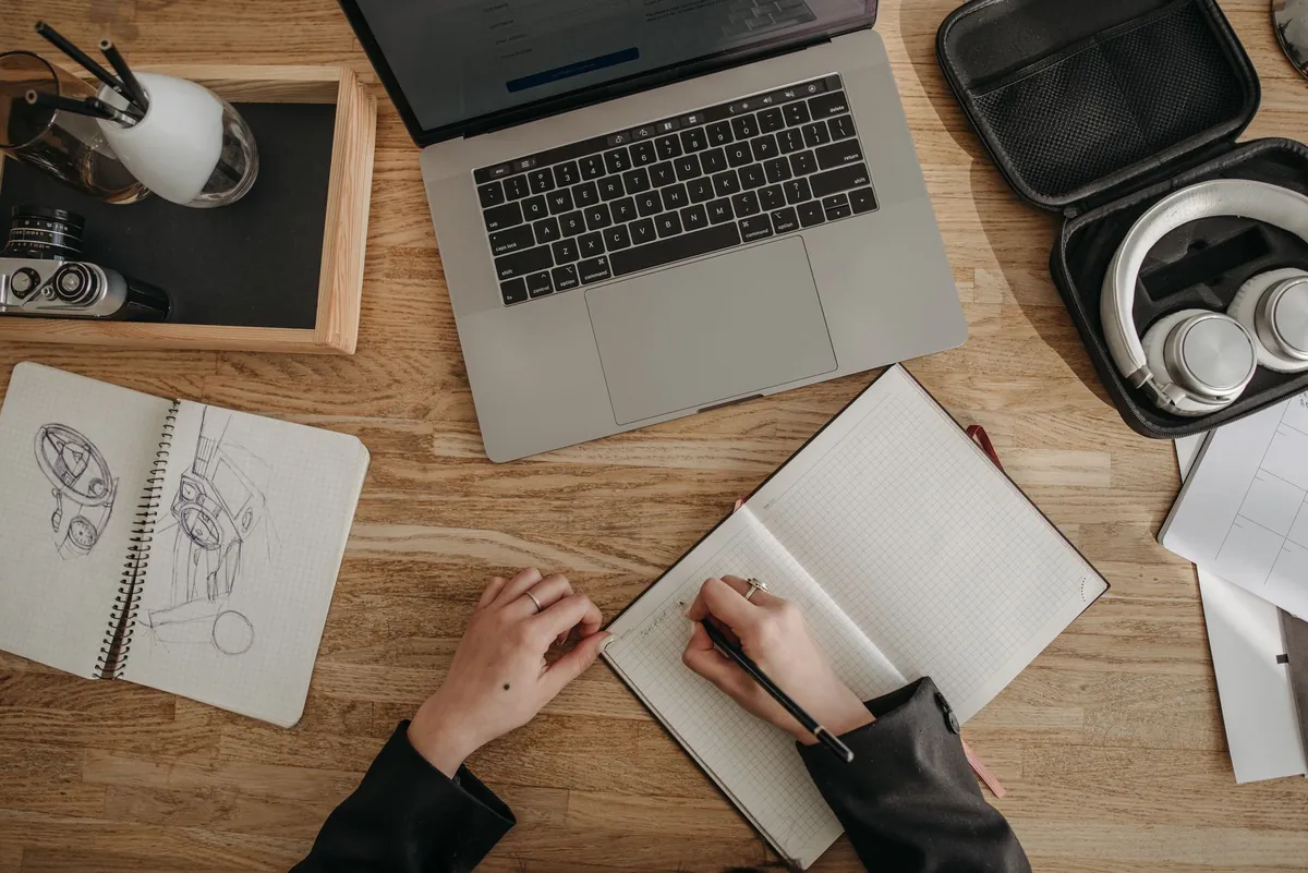 A workspace seen from above with a laptop, sketchbooks and pens spread across a wooden table