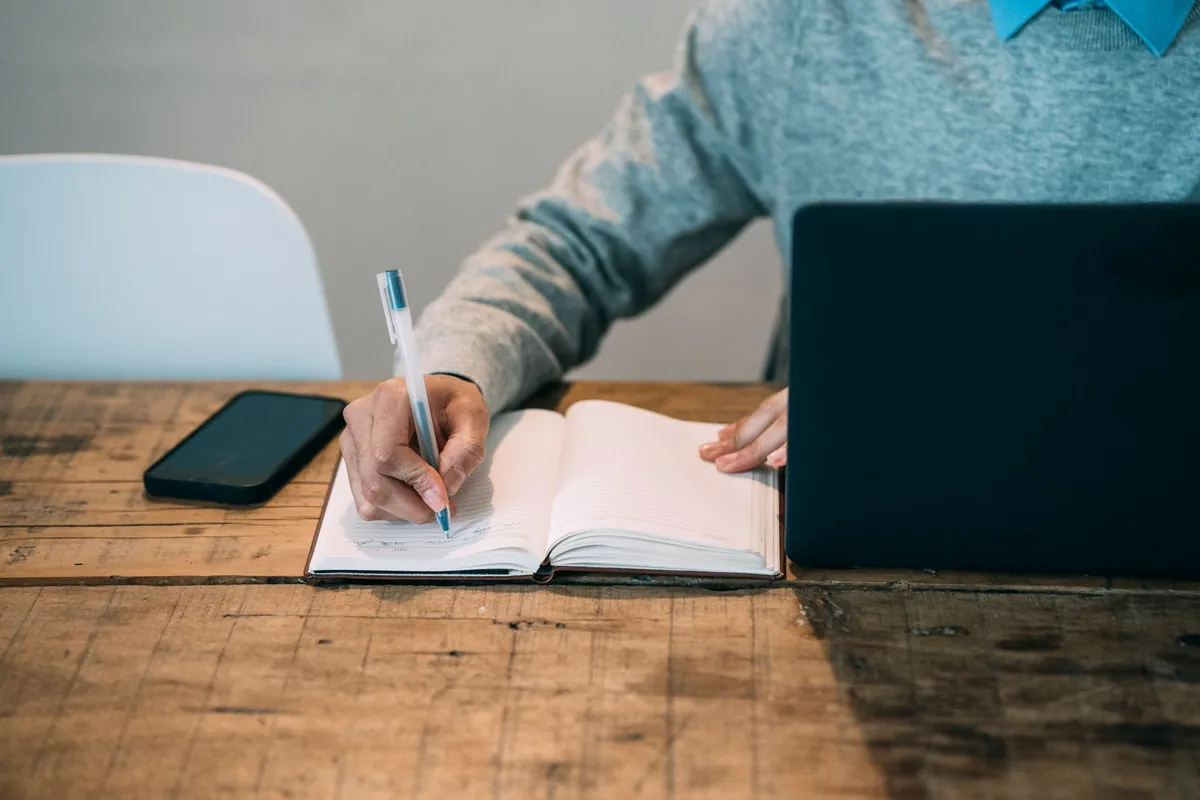 A person writing in a notebook at a wooden table beside a small laptop