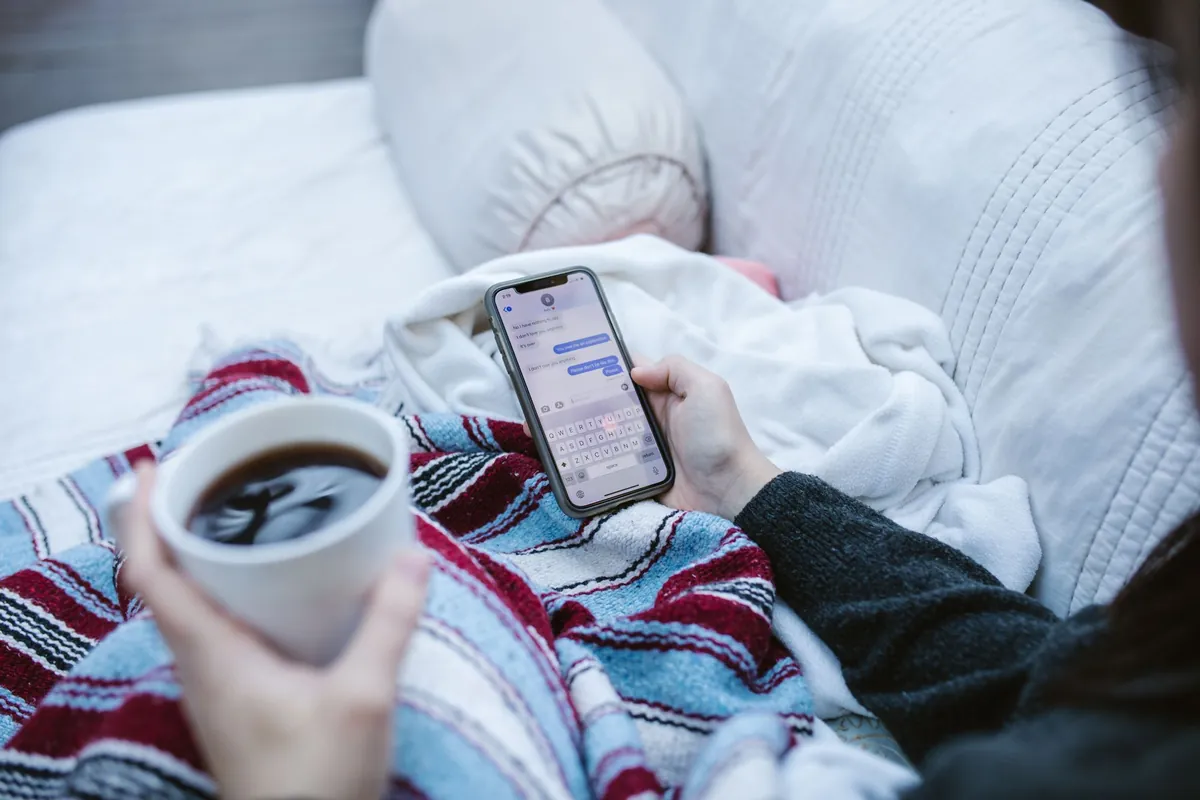 A person holding a phone showing a message thread, coffee in the other hand, reading at home