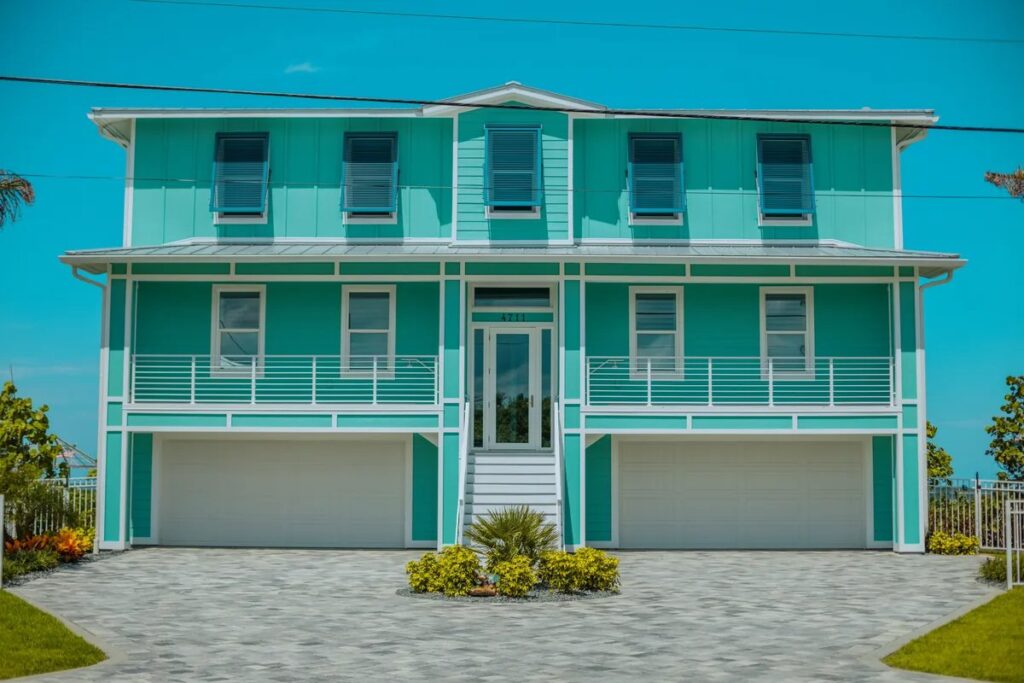 Two-story turquoise coastal house with blue storm shutters, white balconies, and a paved driveway under a clear sky