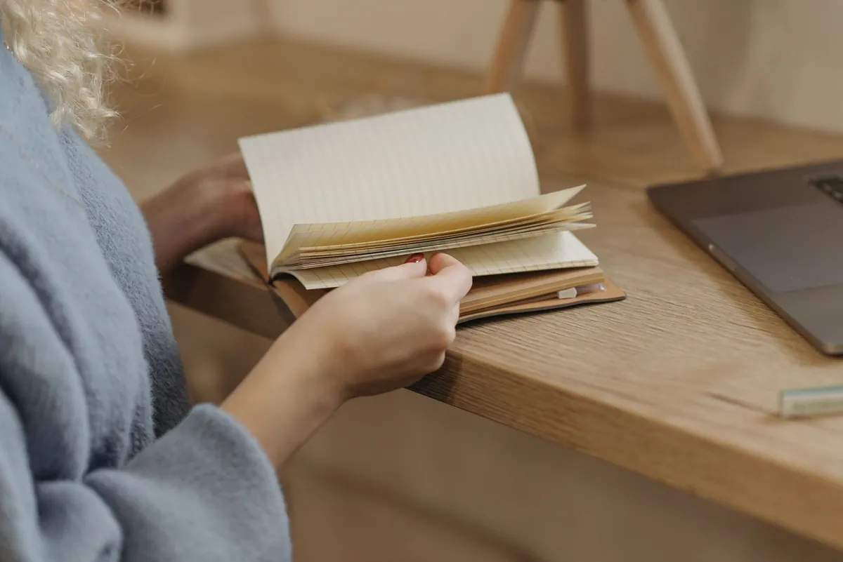 A person turning the pages of a notebook beside a laptop on a wooden desk