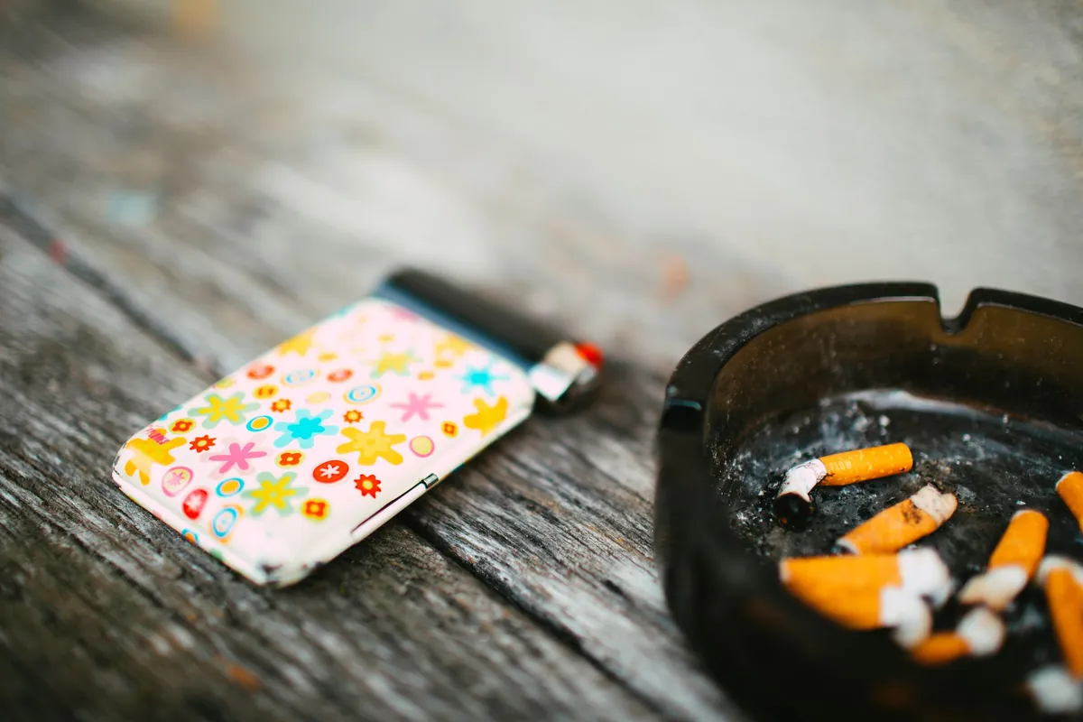 A black ashtray full of cigarette butts beside a flowered lighter on a weathered table