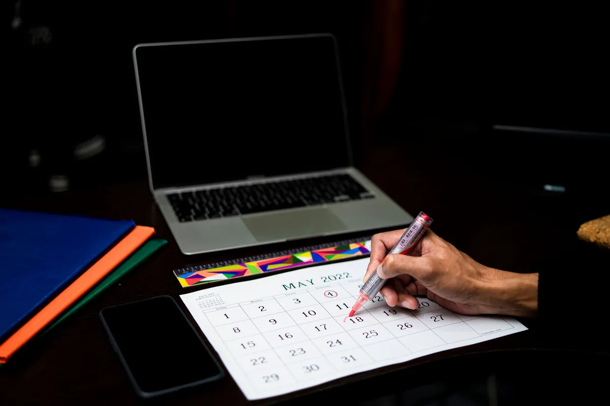 A wall calendar and an open laptop on a dark desk, with a hand writing