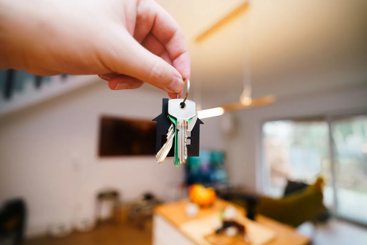 A hand holding a set of keys with a house keyring inside a bright, furnished short-term rental