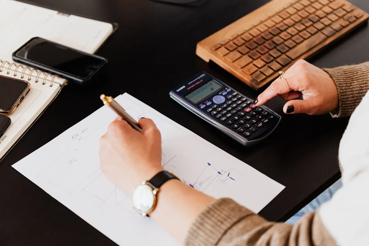 A hand writing figures on a sheet of paper beside a calculator and a phone