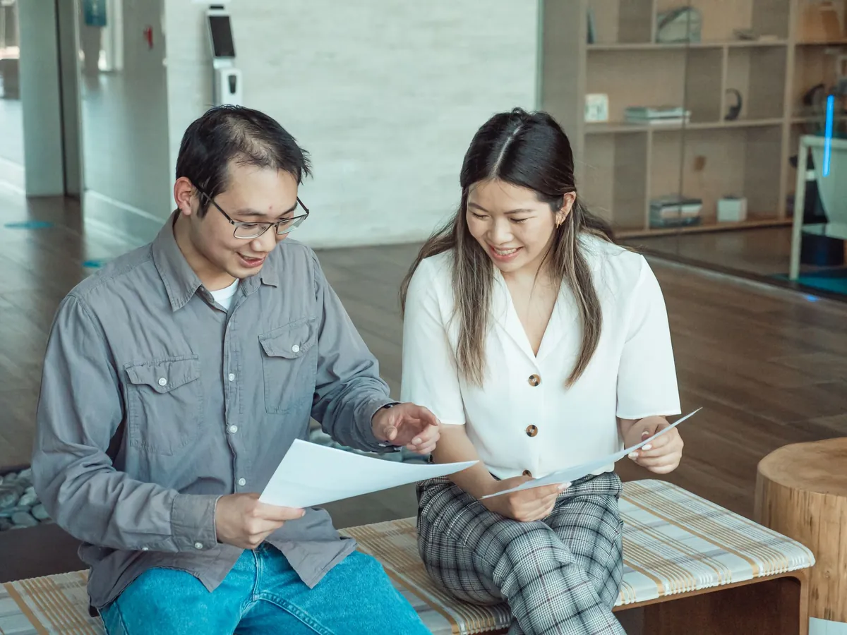 Two people sitting side by side, each holding a sheet of paper, comparing them