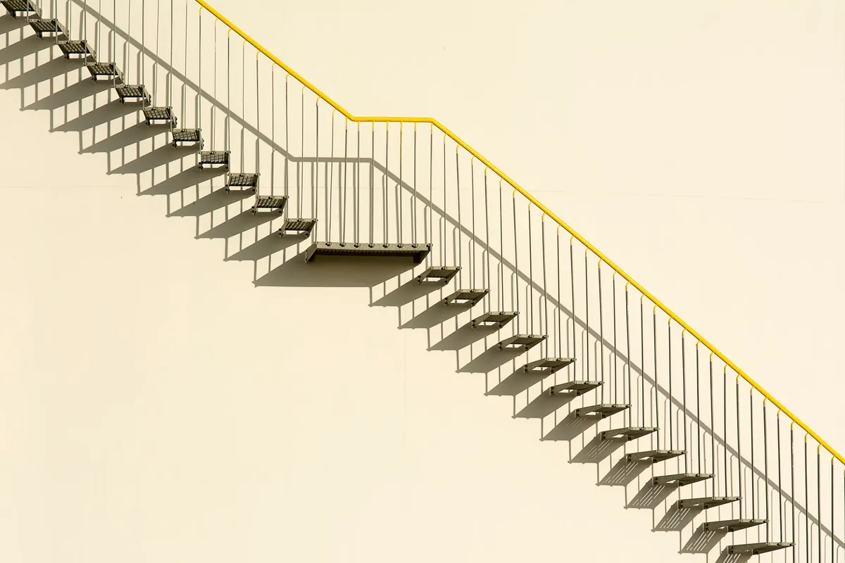 An external metal staircase shot square-on from the side, its steps and yellow handrail climbing diagonally across a plain cream wall with the railing shadow beside it.