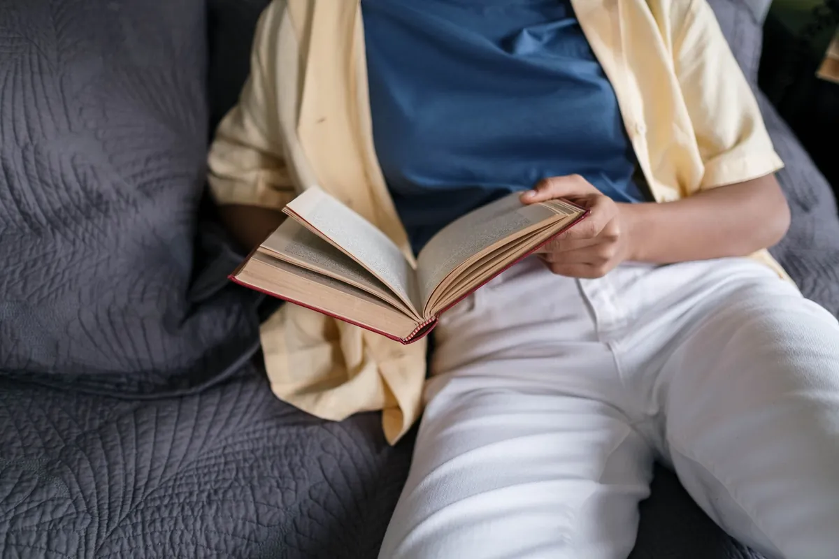 A person sitting back on a grey quilted sofa in a yellow shirt and white trousers, holding an open hardback book on their lap, framed from the neck down.