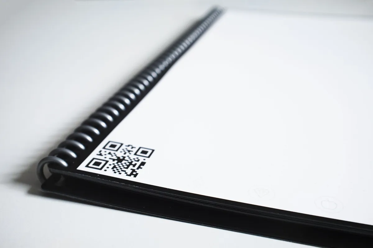 The top corner of a black spiral-bound notebook on a white surface, with a printed QR code in the corner of the blank first page.