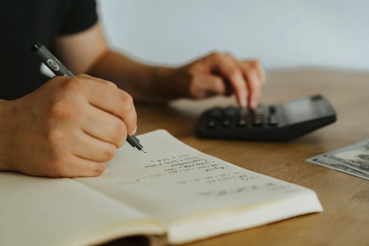 A hand writing a column of figures in a notebook beside a calculator